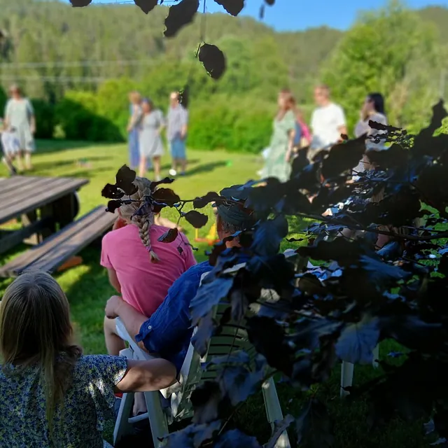 Folk sitter på stoler i skygge og ser på gruppe som danser på gressplen i solskinn.