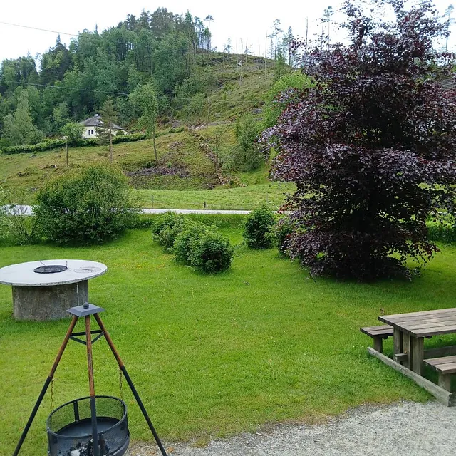 Uteområde med bålpanne, benker og grønt gress, med åsside og hus i bakgrunnen.