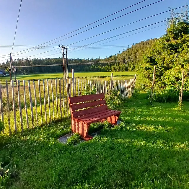 Rød trebenk ved gjerde med utsikt over grønne jorder og skog i kveldssol.