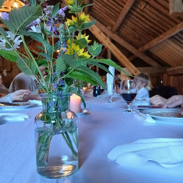 Nærbilde av markblomster i vase på langbord med hvit duk i rustikk låve.